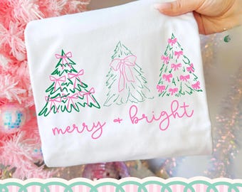 May include: A white t-shirt featuring a graphic of three stylized Christmas trees in green and pink. The trees are decorated with pink bows. Below the trees, the text merry + bright is printed in a pink cursive font.