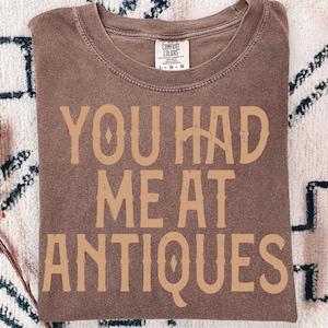 May include: A brown Comfort Colors t-shirt with the text "YOU HAD ME AT ANTIQUES" in a tan, vintage-style font. The shirt is folded and has a tag in the upper center.