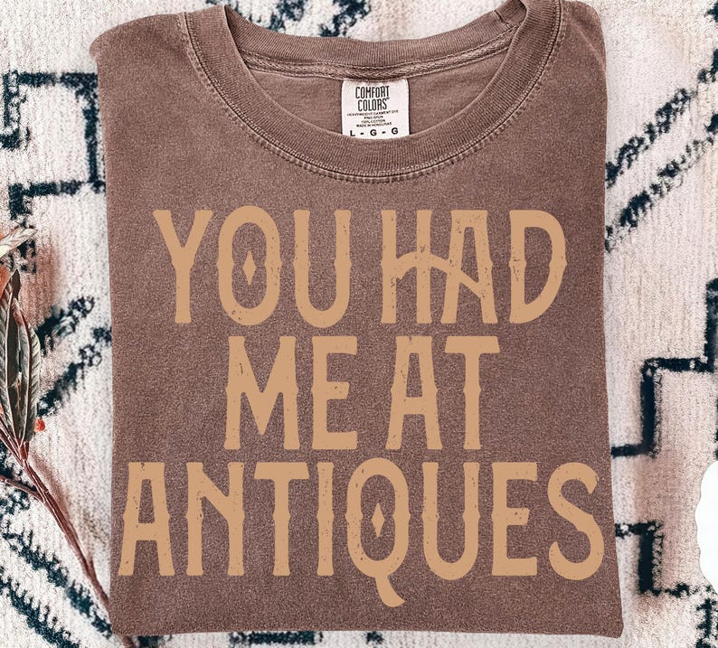 May include: A brown Comfort Colors t-shirt with the text "YOU HAD ME AT ANTIQUES" in a tan, vintage-style font. The shirt is folded and has a tag in the upper center.