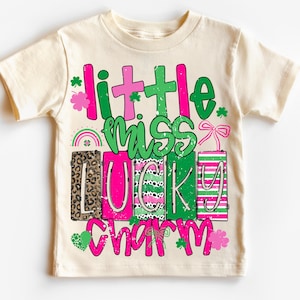 Puede incluir: Camiseta crema para niños con la frase "Little Miss Lucky Charm" en rosa y verde. El diseño incluye tréboles, un arcoíris y un lazo. Las letras tienen un efecto desgastado.
