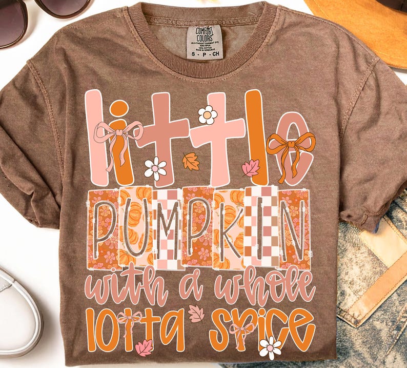 Op de afbeelding: Een bruin t-shirt met korte mouwen en een grafische print met de tekst "little PUMPKIN with a whole lotta SPICE". Het ontwerp bevat roze en oranje letters, kleine witte bloemen, oranje herfstbladeren en oranje strikjes op de letters.