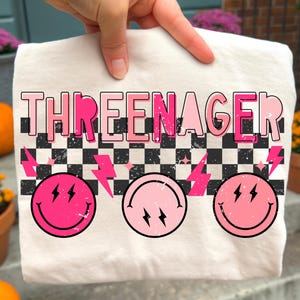 May include: Cream-colored t-shirt with the text "THREENAGER" in pink, above a black and white checkered pattern and three pink smiley faces with lightning bolts. The shirt is held up by a hand.