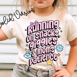 Op de afbeelding: Een wit T-shirt met een roze afbeelding waarop staat "Running on snacks, giggles & moms patience". De afbeelding is versierd met kleurrijke bloemen en sterren.