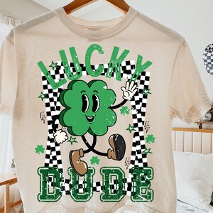 May include: Beige t-shirt with a green cartoon clover design. The clover has a smiling face and is waving. The words "LUCKY DUDE" are printed in green, with a black and white checkered border.