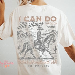 May include: A white t-shirt with a graphic print of a cowboy riding a horse in a desert landscape. The text "I can do all things through Christ which strengtheneth me Philippians 4:13" is printed on the shirt.