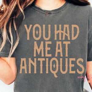May include: Dark gray t-shirt with the text "YOU HAD ME AT ANTIQUES" in a vintage-style, tan font. The shirt has a slightly faded appearance, suggesting a vintage or retro aesthetic. The design is centered on the chest.
