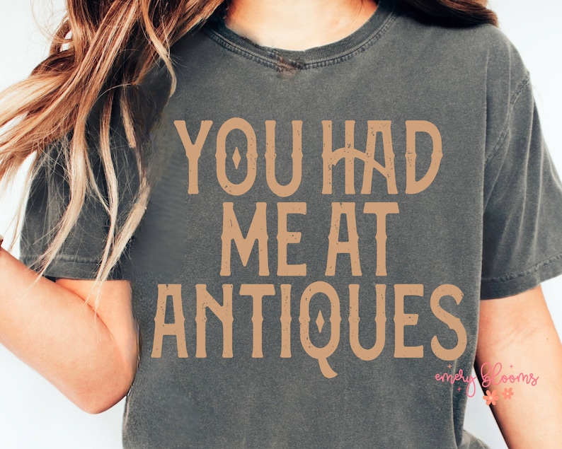 May include: Dark gray t-shirt with the text "YOU HAD ME AT ANTIQUES" in a vintage-style, tan font. The shirt has a slightly faded appearance, suggesting a vintage or retro aesthetic. The design is centered on the chest.