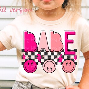 May include: A white t-shirt with the word "BABE" in pink and white letters. The letters are outlined in black and have a checkered pattern behind them. There are three pink smiley faces with lightning bolts below the word.