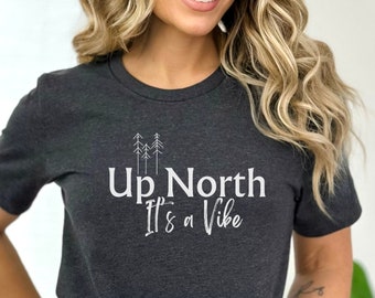 Puede incluir: Una camiseta gris oscuro con un gráfico blanco que dice "Up North It's a Vibe" con una pequeña imagen de pinos.