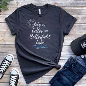 Puede incluir: Una camiseta gris oscuro con el texto "Life is better on Butterfield Lake" en letras blancas. La camiseta presenta un gráfico de sol y olas. La camiseta está colocada sobre una superficie de madera con un par de zapatillas negras, unos vaqueros azules y una gorra de béisbol negra.