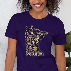 May include: A purple t-shirt with a gold outline of the state of Minnesota. The design includes phrases such as "Skol," "Minnesota Nice," and "Uff-da." The shirt is a short-sleeved crew neck.