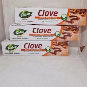 Peut inclure: Trois boîtes rectangulaires de dentifrice Dabur Clove Cavity Protection empilées verticalement. L'emballage blanc présente le nom de la marque, un logo feuille verte, une image de clous de girofle sur bois et 0% de fluor ajouté.