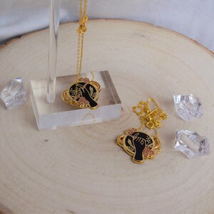 Pode incluir: Dois colares de ouro com pingentes pretos e dourados. Os pingentes apresentam uma mão segurando um círculo com detalhes florais. Os colares estão sobre uma superfície de madeira.