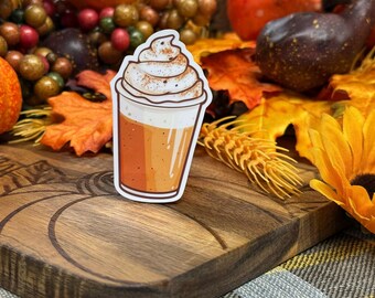Op de afbeelding: Een sticker met een herfstthema van een pumpkin spice latte met slagroom en kaneel erop. De sticker zit op een houten oppervlak met herfstdecoraties.