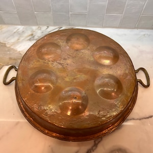 May include: A copper escargot pan with six circular indentations. The pan has a round shape with two brass handles. The copper has a warm, aged patina, with variations in color from reddish-brown to golden hues.