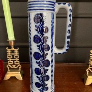 May include: A tall, white ceramic beer stein with a blue and white floral design. The stein has a handle and a wide base.