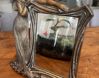 Op de afbeelding: Een vintage messing spiegel met een decoratief frame met een gesculpteerd vrouw in een vloeiende jurk. De spiegel heeft een gefacetteerde rand en een helder glazen oppervlak.