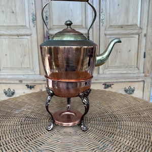 May include: Antique copper and brass tea kettle with a decorative stand. The kettle features a copper body, brass spout, lid, and handle. The stand has ornate legs and a round base. A vintage kitchenware item.