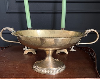 May include: A gold-toned metal bowl with two handles sits on a wooden surface. The bowl is on a pedestal and has a decorative design around the rim. Two green candles are in the background.