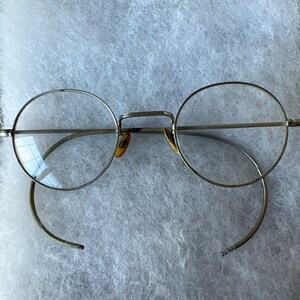 May include: Silver round glasses with a thin metal frame and clear lenses. The glasses are lying on a white fuzzy surface.