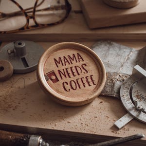 Puede incluir: Un posavasos de madera con la inscripción grabada "MAMA NEEDS COFFEE" y un dibujo de una taza de café. El posavasos es de color marrón claro y está sobre una superficie de madera con herramientas de carpintería.
