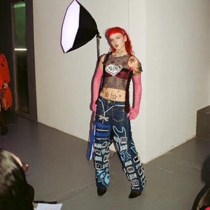 May include: A person with pink hair and pink gloves is wearing a black mesh top with a red heart that says "MAMA" on it. They are also wearing blue jeans with blue and white patches and a dragonfly charm on the belt loop.