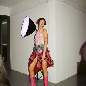 以下が含まれることがあります： ピンクと白のクロップトップ、赤と黒のチェック柄のスカート、ピンクのブーツを履いた女性。彼女は左手に大きな白いライトリフレクターを持っています。