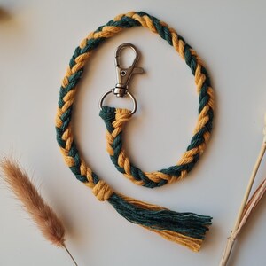 May include: Braided lanyard in green and gold. The lanyard features a metal clasp and a tassel at the end. The lanyard is made of a soft, woven material. Decorative dried flowers and plants are in the background.