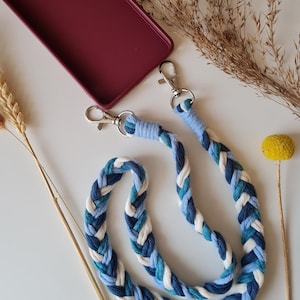 Puede incluir: Una correa de teléfono trenzada con hilo de color azul, blanco y turquesa. La correa tiene dos cierres plateados para sujetar a una funda de teléfono.