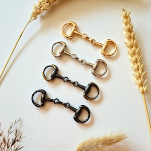 May include: Four metal horse bit charms in gold, silver, and black, arranged on a white surface with dried wheat stalks. The bits have a classic design with D-rings and a central link.