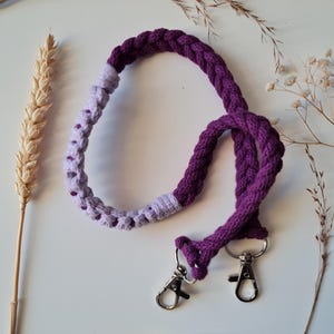 May include: A handmade purple and lavender braided lanyard with silver-colored metal clasps. The lanyard is made of thick, soft rope and is designed to be worn around the neck or shoulder. The background is a white surface with dried plants.