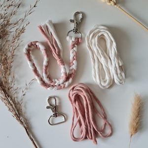 Może przedstawiać: Zestaw breloków i smyczy z makramy w kolorze białym i pudrowym różu. Zestaw zawiera plecioną smycz, oddzielną długość sznurka i dwa srebrne metalowe zapięcia. Suche trawy i kłosy pszenicy ułożone są wokół przedmiotów na białej powierzchni.