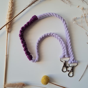 May include: A lavender and purple macrame lanyard with silver-colored swivel clasps. The lanyard is made of braided cord and features a section of deep purple macrame knots. The design is suitable for keys or an ID badge.