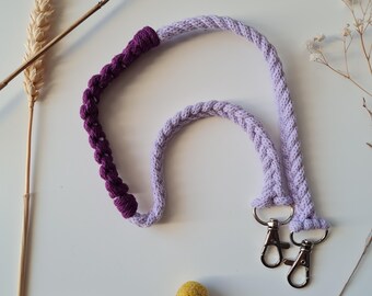 May include: A lavender and purple macrame lanyard with silver-colored swivel clasps. The lanyard is made of braided cord and features a section of deep purple macrame knots. The design is suitable for keys or an ID badge.