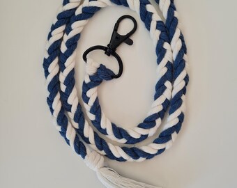 May include: A blue and white braided rope lanyard with a black clasp and a white tassel. The lanyard is made of thick, soft rope and is designed to be worn around the neck. The overall design is nautical.