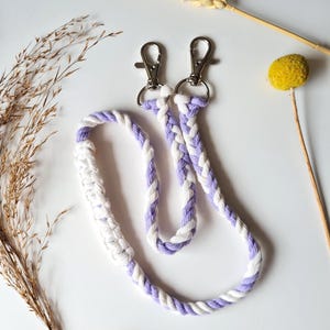 May include: A braided lanyard with a light purple and white rope design. The lanyard has two silver-colored metal clasps at the top. The lanyard is made of a thick, durable rope material. The background includes dried flowers and a white surface.