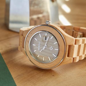 May include: A wooden wristwatch with a light brown band and a silver face. The watch face has the word "BEWELL" printed on it.