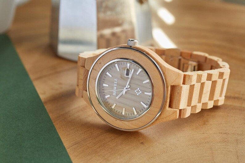 May include: A wooden wristwatch with a light brown band and a silver face. The watch face has the word "BEWELL" printed on it.