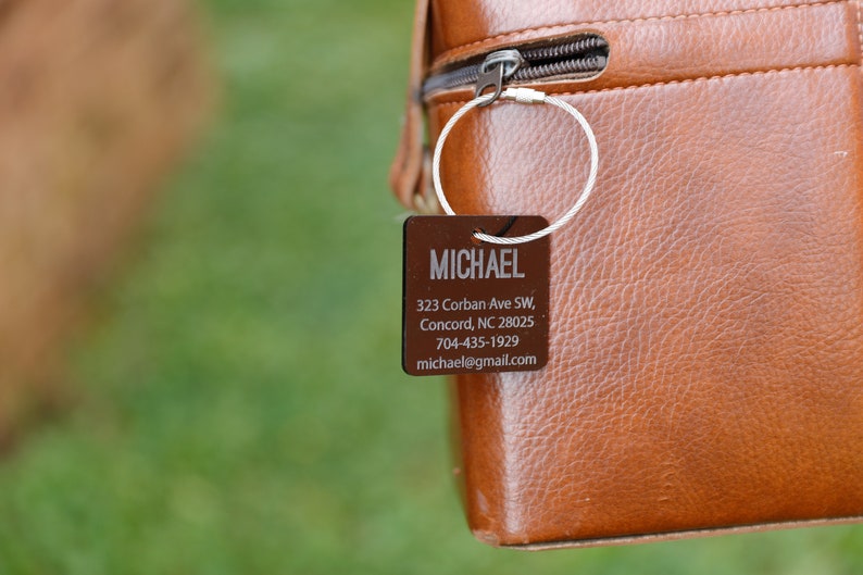 May include: A brown leather bag with a metal ring and a brown rectangular tag attached. The tag has the name "MICHAEL" and contact information: 323 Corban Ave SW, Concord, NC 28025, 704-435-1929, michael@gmail.com.