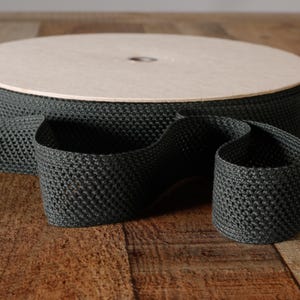 May include: A spool of dark charcoal gray woven webbing tape featuring a distinct textured grid pattern. The sturdy fabric strap is wound tightly around a circular wooden core, with a portion of the material unspooled and resting in front.