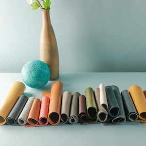 May include: A collection of 15 rolled leather swatches in various colors, including brown, tan, yellow, green, blue, and gray. The swatches are arranged in a row on a light blue surface.
