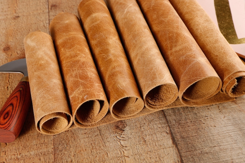 May include: Close-up of six rolled pieces of brown leather on a wooden surface. The leather has a natural, textured look.
