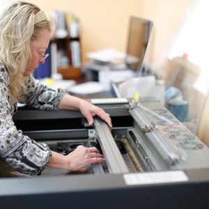 May include: A woman is operating a laser cutting machine. The machine has a clear protective cover and a metal bed. The woman is wearing a patterned blouse and is carefully adjusting the laser head.