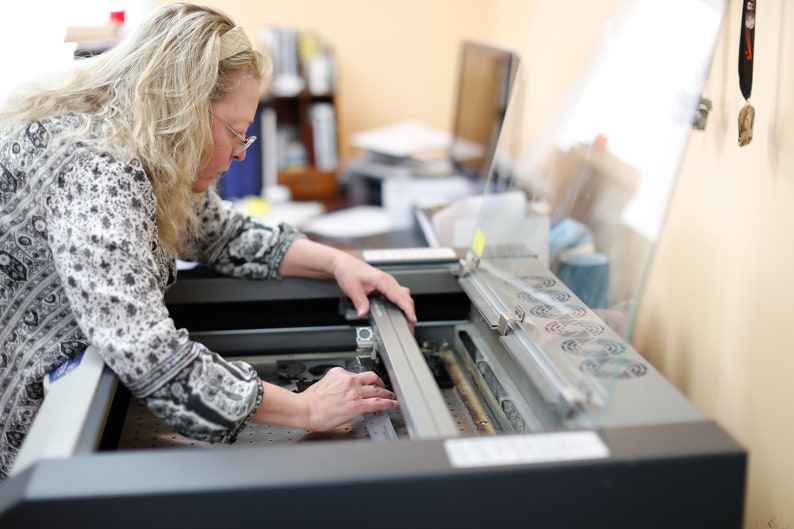 May include: A woman is operating a laser cutting machine. The machine has a clear protective cover and a metal bed. The woman is wearing a patterned blouse and is carefully adjusting the laser head.
