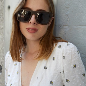 May include: A woman with shoulder-length brown hair wearing a white blouse with a floral pattern and dark brown sunglasses.