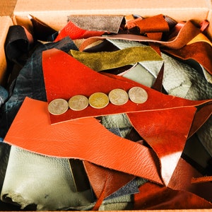 May include: A cardboard box filled with scraps of leather in various colors, including red, brown, green, and blue. A row of coins is placed on top of the leather scraps.