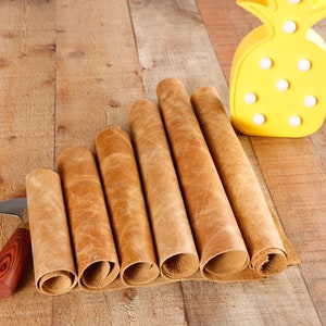 May include: A close-up of six rolls of brown leather on a wooden surface. A small knife with a wooden handle is lying next to the leather rolls. A yellow pineapple-shaped lamp is in the background.