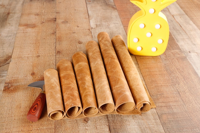 May include: A close-up of six rolls of brown leather on a wooden surface. A small knife with a wooden handle is lying next to the leather rolls. A yellow pineapple-shaped lamp is in the background.