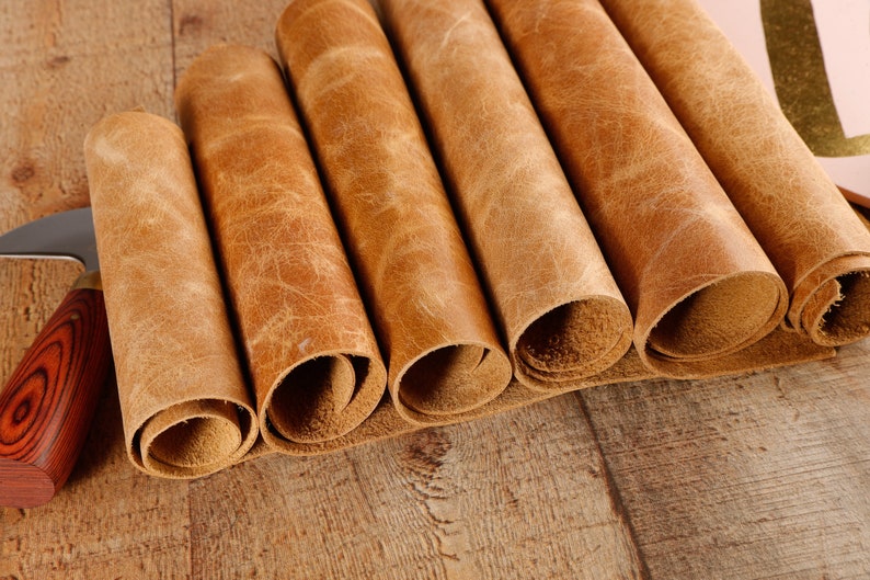 May include: Close-up of six rolled pieces of brown leather on a wooden surface. The leather has a natural, textured look.