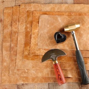 May include: A set of leather crafting tools including a leather cutting knife, a heart-shaped hole punch, and a brass hammer. The tools are laid out on a stack of brown leather sheets.